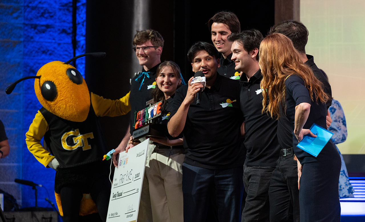 Team Hearo holding a big check and trophy&nbsp;on stage between Buzz and InVenture Prize host, Faith Salie.