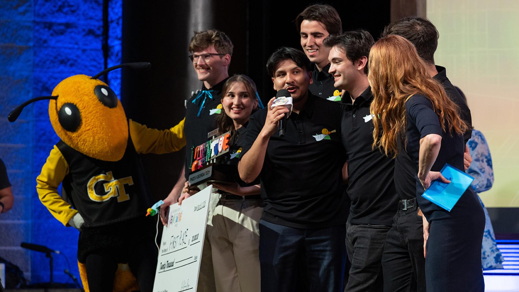 Team Hearo holding a big check and trophy on stage between Buzz and InVenture Prize host, Faith Salie.