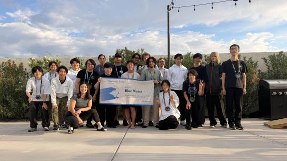 A group of people standing outside holding a banner. The banner says "Blue Water" which is the team's name.