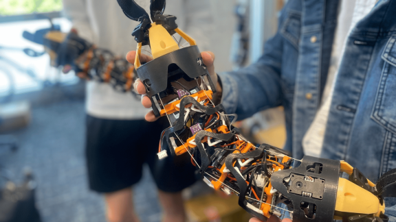 Close-up of a modular, snake-like robotic device with exposed wiring, sensors, and claw-like grippers at both ends being held by two people in a laboratory setting. The flexible robotic structure features black and orange components connected by segmented joints.