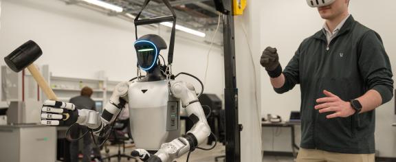 This is an image of someone using VR goggles and a glove to control a humanoid robot holding a rubber mallet.