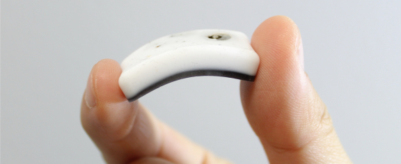 Close-up of a flexible white wearable sensor device held between two fingers, showing its curved shape against a plain light-gray background.