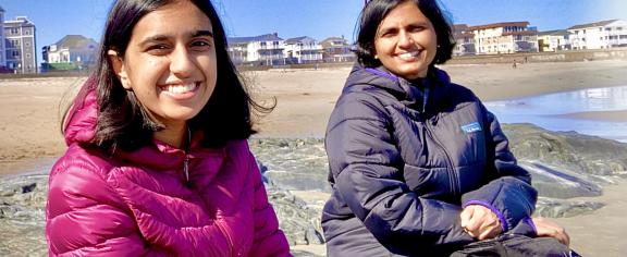 Two smiling researchers wearing jackets sit on rocks at a sunny beach with shoreline homes and the ocean in the background.