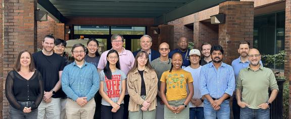 A group of students, researchers, and staff stand together for a group photo outside a brick building beneath a covered walkway.