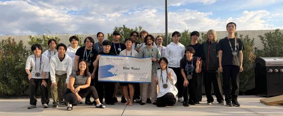 A group of people standing outside holding a banner. The banner says "Blue Water" which is the team's name.