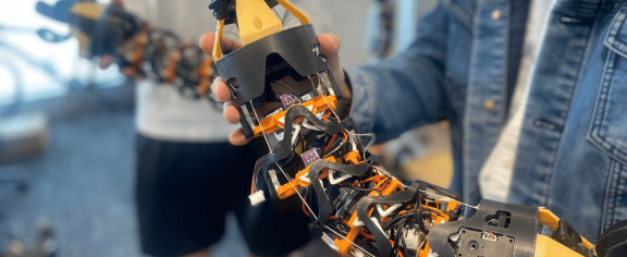 Close-up of a modular, snake-like robotic device with exposed wiring, sensors, and claw-like grippers at both ends being held by two people in a laboratory setting. The flexible robotic structure features black and orange components connected by segmented joints.