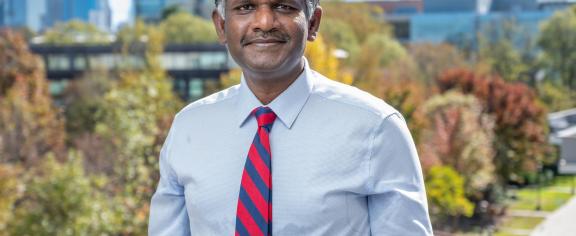 Photo of a man of south Asian descent with short dark hair and a smile. He is wearing a light blue button-down shirt and a red and royal blue striped tie. Behind him is the Atlanta skyline.