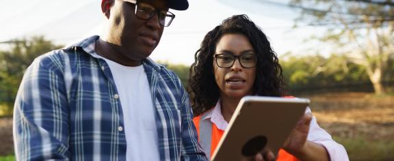 A farmer holds a tablet while a woman to his right assists him