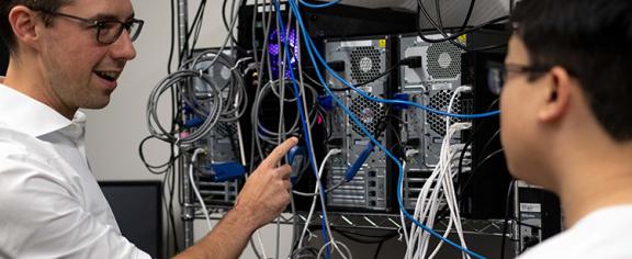 A man points to a rack of CPUs. He is talking to a student who is slighlty out of focus. 