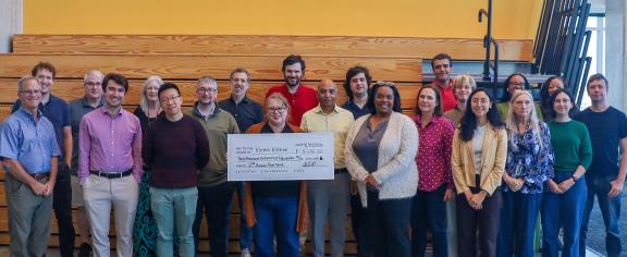 A group photo of more than a dozen people. A woman in the center is holding a large check for Klemis Kitchen, Georgia Tech's on-campus food pantry. 