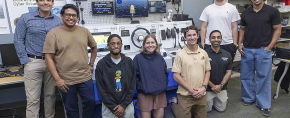 A group photo of students and faculty standing around lab equipment found on a ship. 