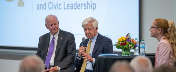 A man speaks into a microphone in front of a banner that reads "Institute for Technology and Civic Leadership" as others look on.