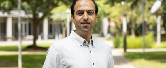 Portrait of an individual standing on a paved campus walkway, wearing a light-colored button-down shirt. Trees, landscaped green spaces, and campus buildings appear in the softly blurred background, with daylight illuminating the outdoor scene. The image is framed from the waist up, with the individual centered in the foreground.