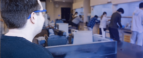 A student wearing safety glasses works in the foreground of a robotics lab as several other students work at equipment in the background.