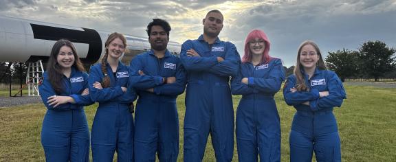 Five people wearing matching blue flight suits stand with arms crossed in a grassy field beside a large rocket display. Sunlight breaks through clouds overhead in the background.
