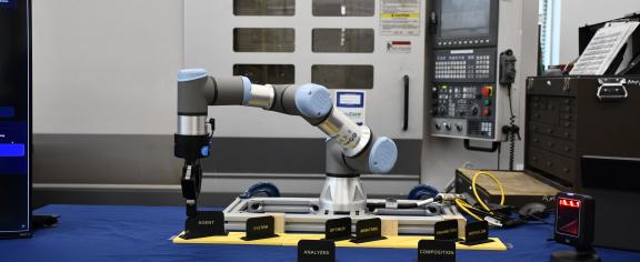 A collaborative robotic arm positioned at a workstation in front of an Okuma CNC machine, surrounded by monitoring equipment and workflow labels.